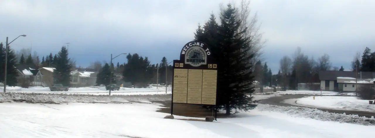 The Welcome to Breton sign beside snowy lawns and spruce trees on a winter day